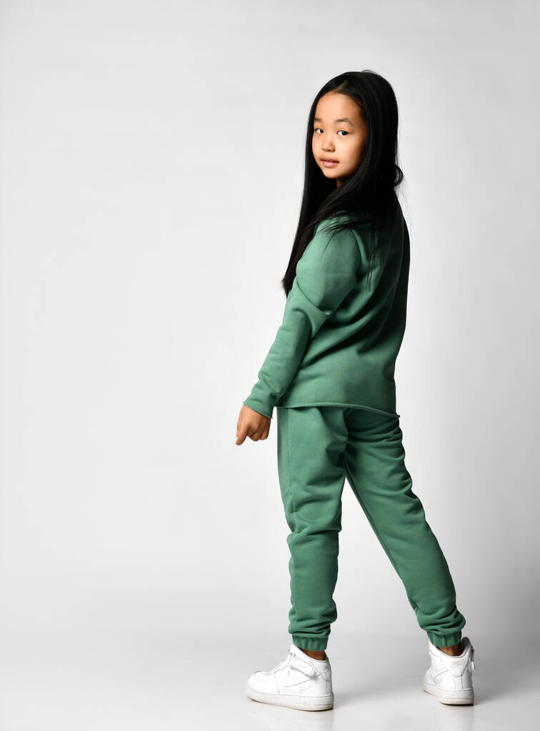 kleines asiatisches Mädchen im trendigen Trainingsanzug, das Spaß auf weißem Hintergrund hat. Ein Kind mit emotionalem Gesichtsausdruck steht halbseitig und posiert. - Foto, Bild