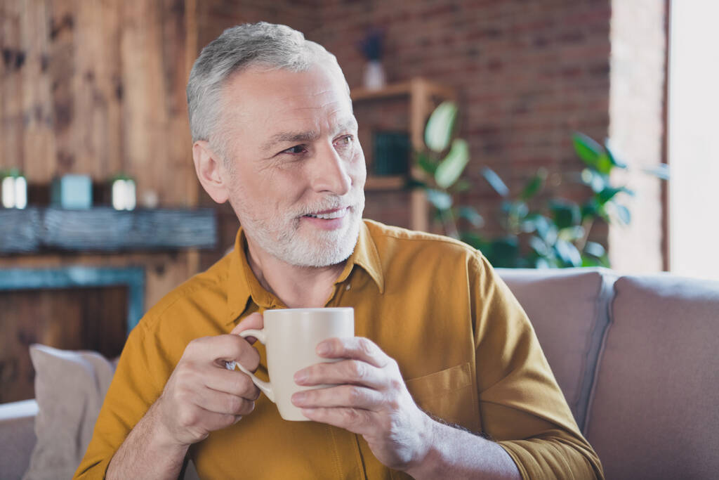 Portret van vrolijke oude man zitten op de bank handen houden koffie kijken geïnteresseerd ver tv tand glimlach genieten weekend binnen - Foto, afbeelding