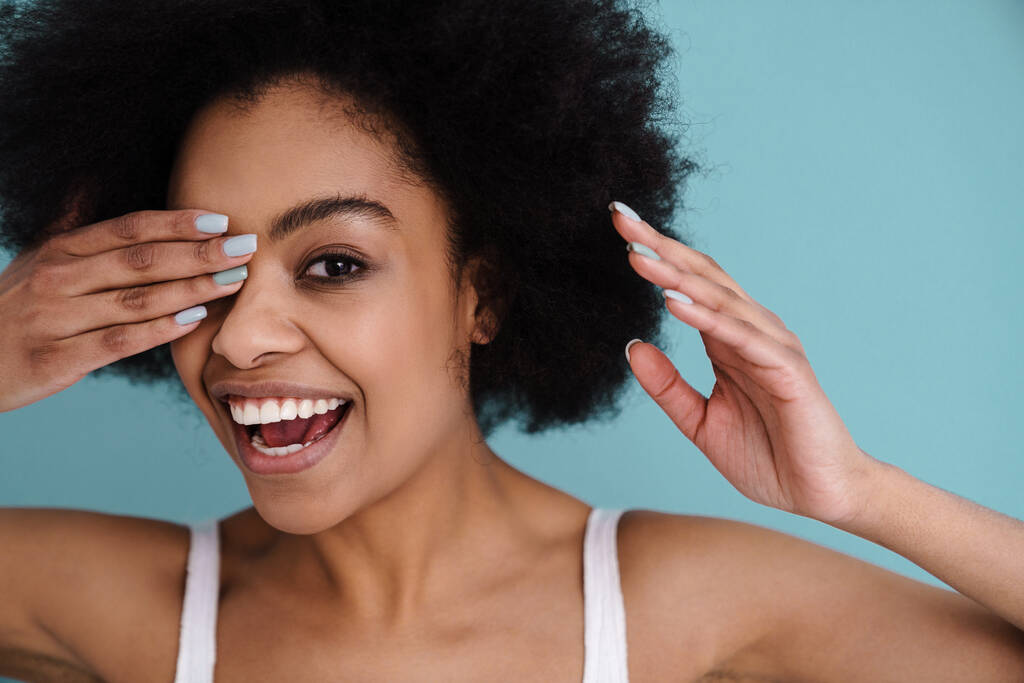Divertente africano americano ragazza sorridente mentre coprendo il suo occhio isolato su sfondo blu - Foto, immagini