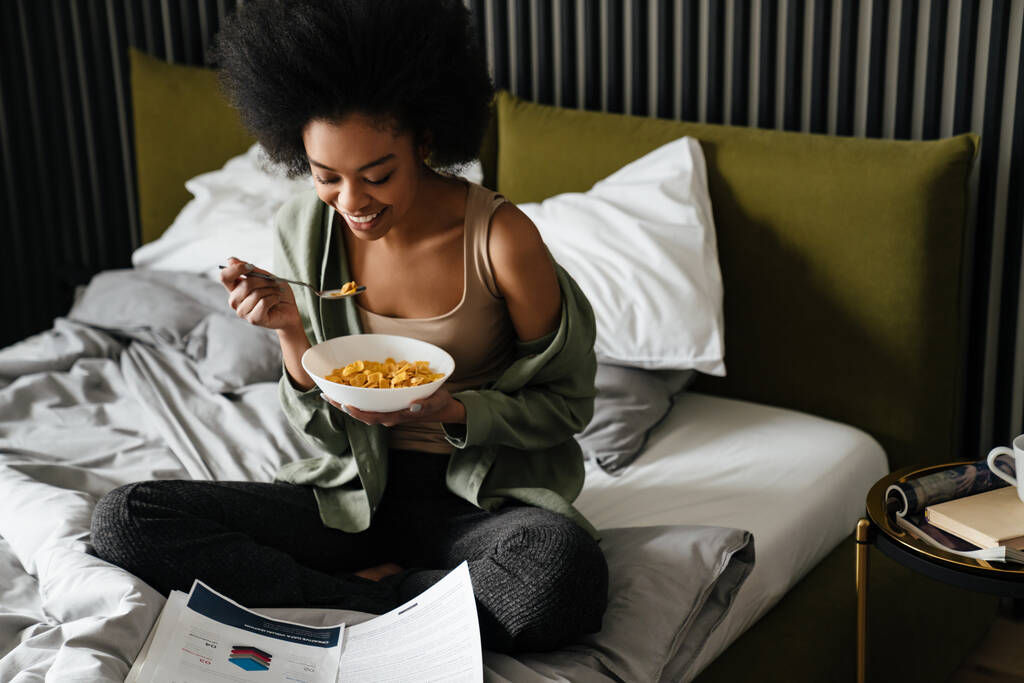 gelukkig afrikaans amerikaans meisje onderzoeken documenten terwijl het eten van granen in bed thuis - Foto, afbeelding