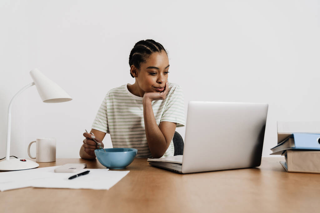 Μαύρη νεαρή γυναίκα τρώει μεσημεριανό ενώ δουλεύει με φορητό υπολογιστή στο γραφείο - Φωτογραφία, εικόνα