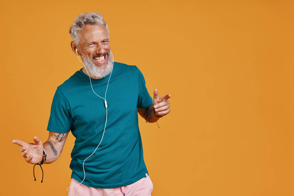 Homme âgé heureux écoutant de la musique et souriant tout en se tenant debout sur fond orange - Photo, image