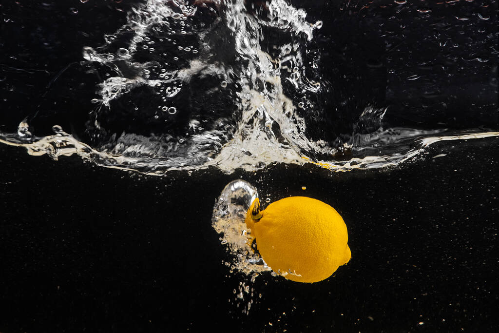Świeżo żółta cytryna wpadająca do wody z odrobiną wody i pęcherzyków powietrza, odizolowana na czarnym tle. - Zdjęcie, obraz