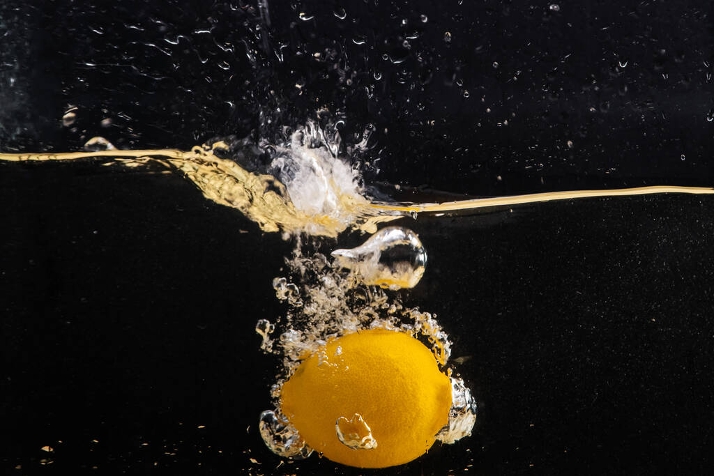 Свежий желтый лимон падает в воду с брызгами воды и воздуха пузырьки, изолированные на черном фоне. - Фото, изображение