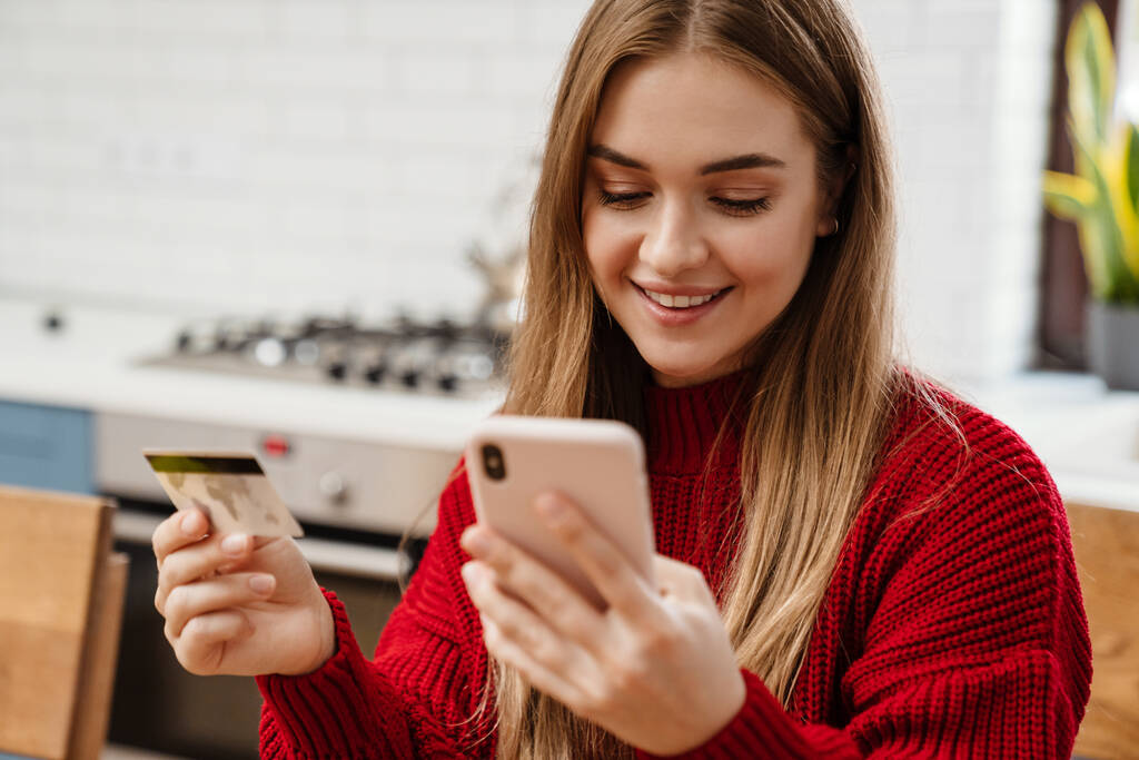Mujer joven bastante alegre desayunando mientras está sentado en la mesa de la cocina, utilizando el teléfono móvil y la tarjeta de crédito para ir de compras en línea - Foto, imagen