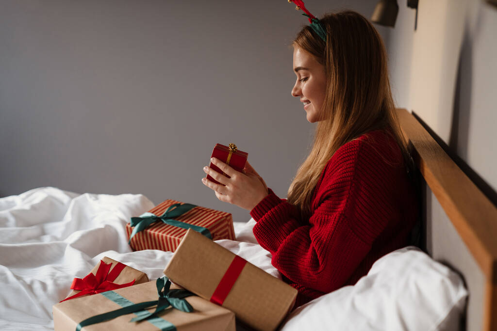 Szczęśliwa młoda dziewczyna w czerwonej piżamie otwierając prezenty świąteczne siedząc w łóżku - Zdjęcie, obraz
