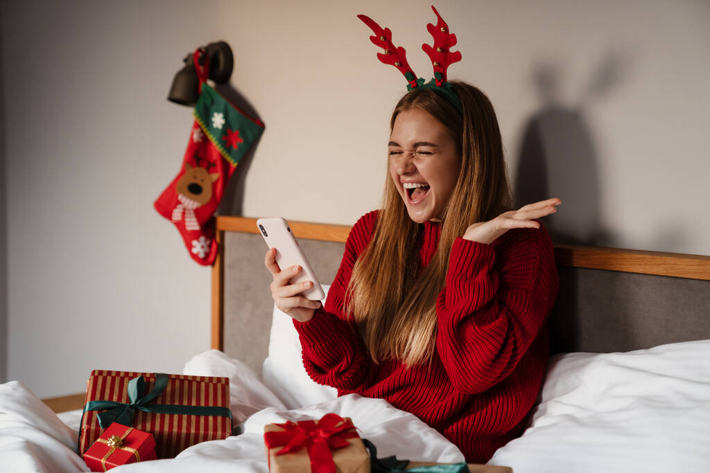 Mujer joven y feliz vistiendo pijamas rojos acostados en la cama con regalos de Navidad, usando el teléfono móvil, celebrando - Foto, imagen