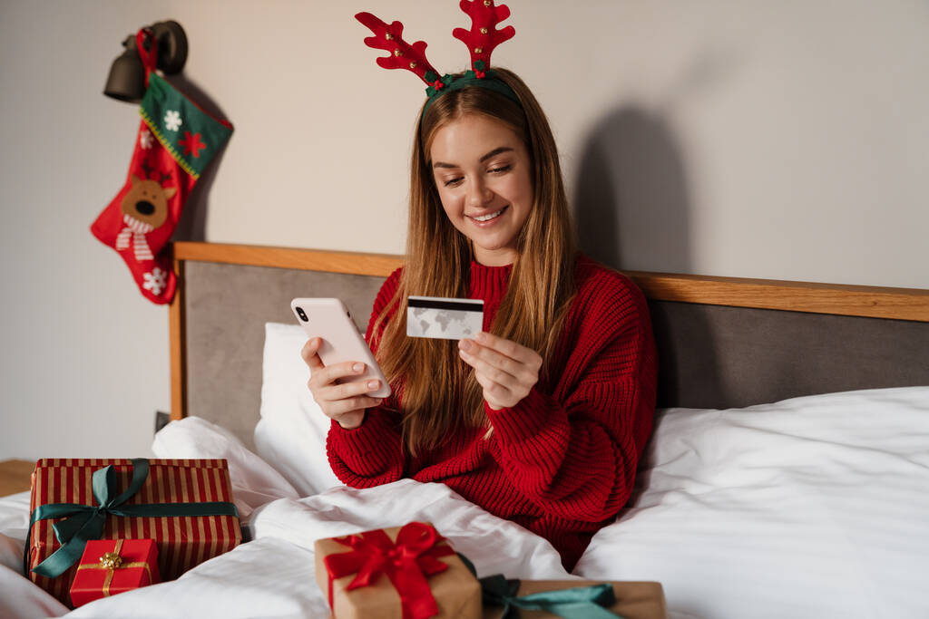 Happy young woman wearing red pajamas laying in bed with christmas presents, using mobile phone and credit card for shopping online - Photo, Image