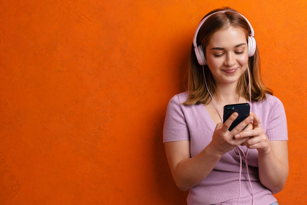 Jeune gingembre femme heureuse écoutant de la musique avec écouteurs et téléphone cellulaire isolé sur un mur orange - Photo, image