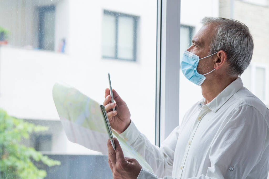 человек в медицинской маске с картой мира в руке и в другом мобильный телефон, концепция путешествия в новой норме - Фото, изображение