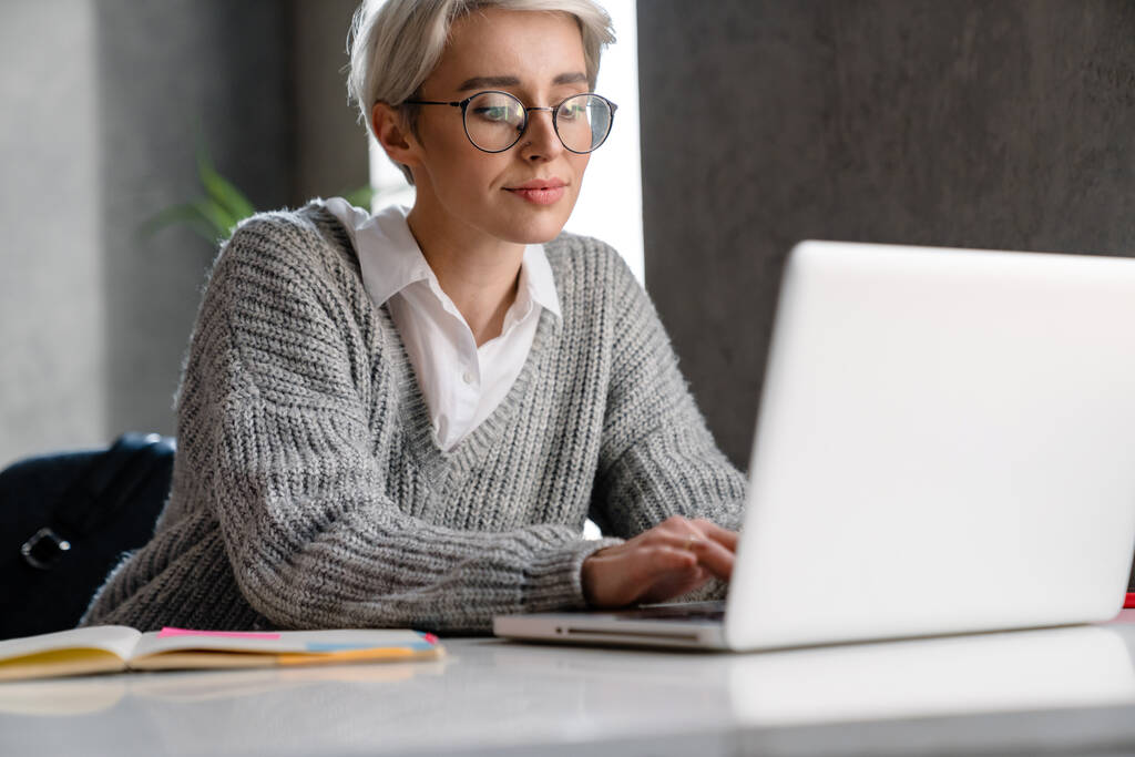 Femme focalisée aux cheveux blancs travaillant avec un ordinateur portable alors qu'elle était assise à table au bureau - Photo, image