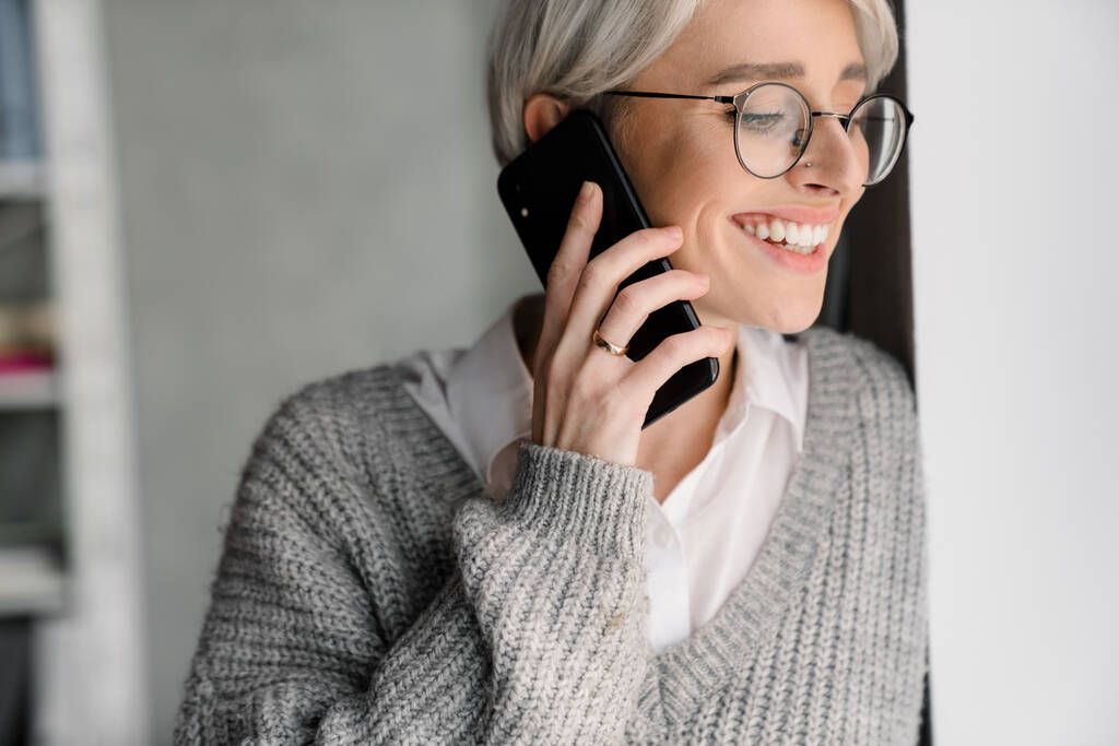 Witharige lachende vrouw in gesprek op mobiele telefoon terwijl ze op kantoor staat - Foto, afbeelding