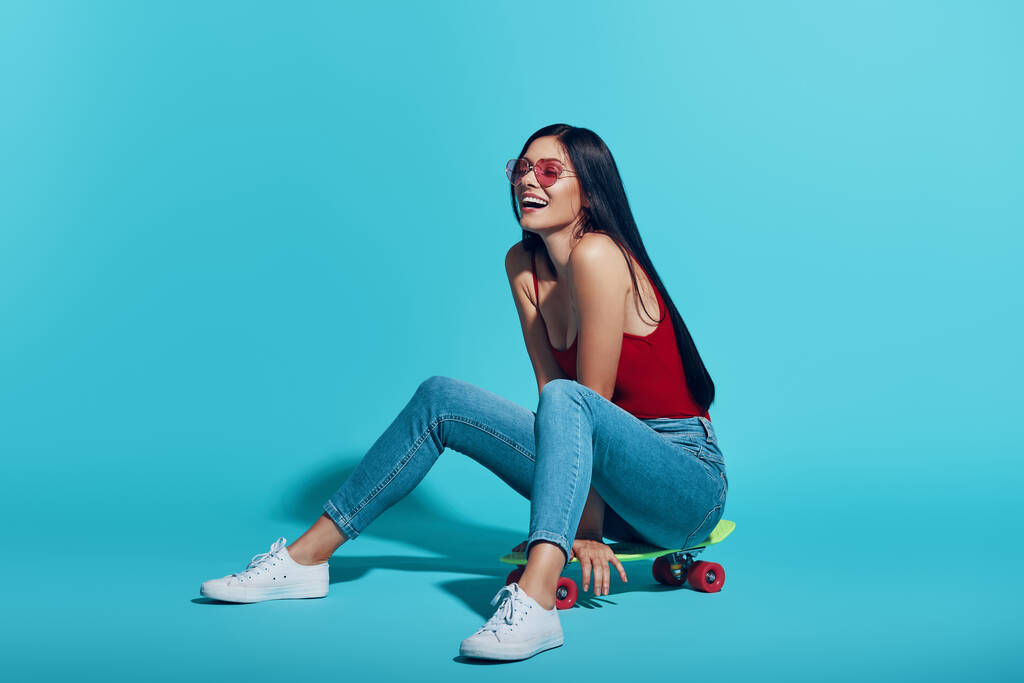 Attraktive junge Frau lächelt auf Skateboard vor blauem Hintergrund - Foto, Bild
