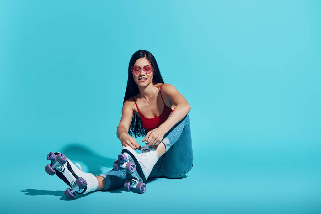 Attractive young woman tying roller skate and smiling while sitting against blue background - Photo, Image