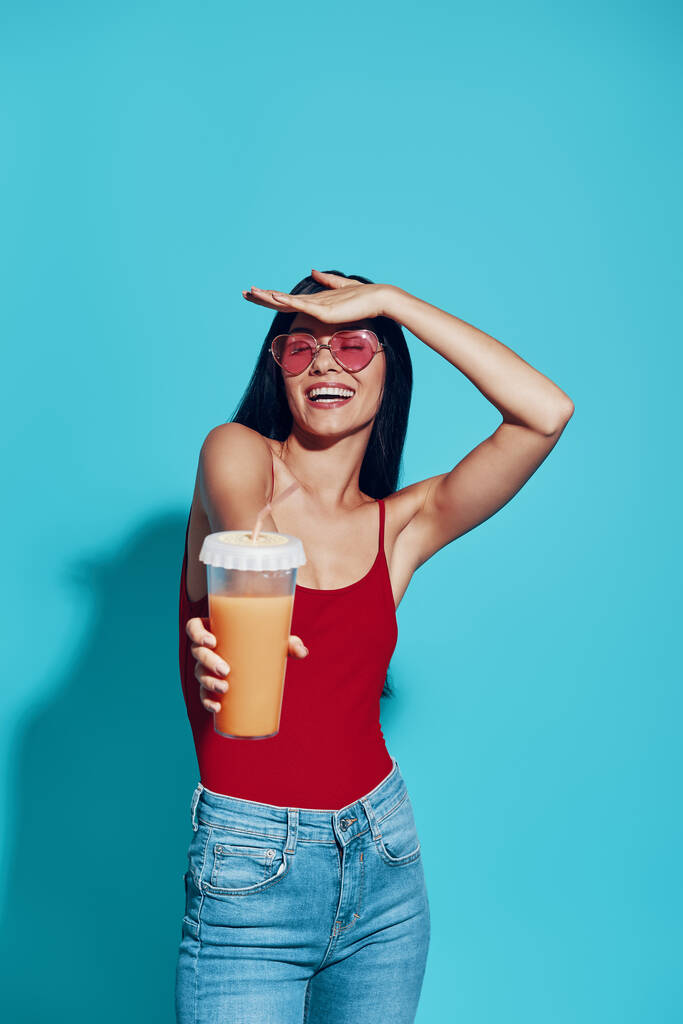 Привлекательная молодая женщина улыбается и дает вам коктейль, стоя на синем фоне - Фото, изображение