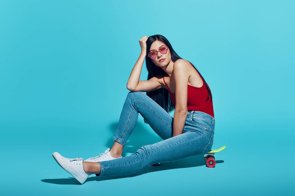 Attraktive junge Frau blickt auf Kamera, während sie auf Skateboard vor blauem Hintergrund sitzt - Foto, Bild