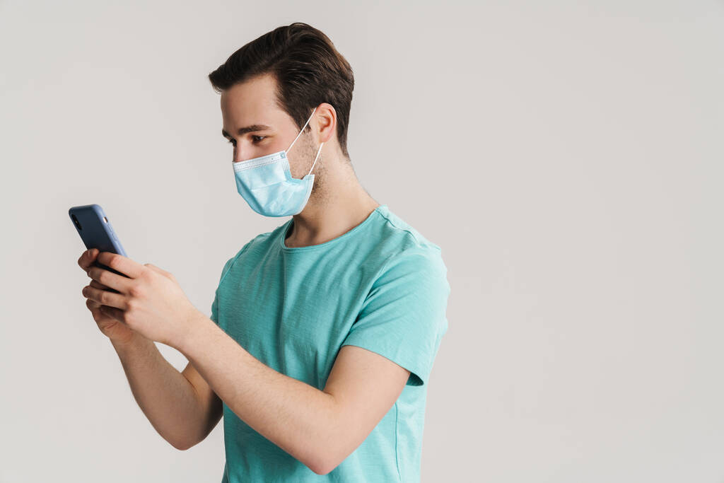 Focused handsome guy in protective mask using mobile phone isolated over white wall - Photo, Image