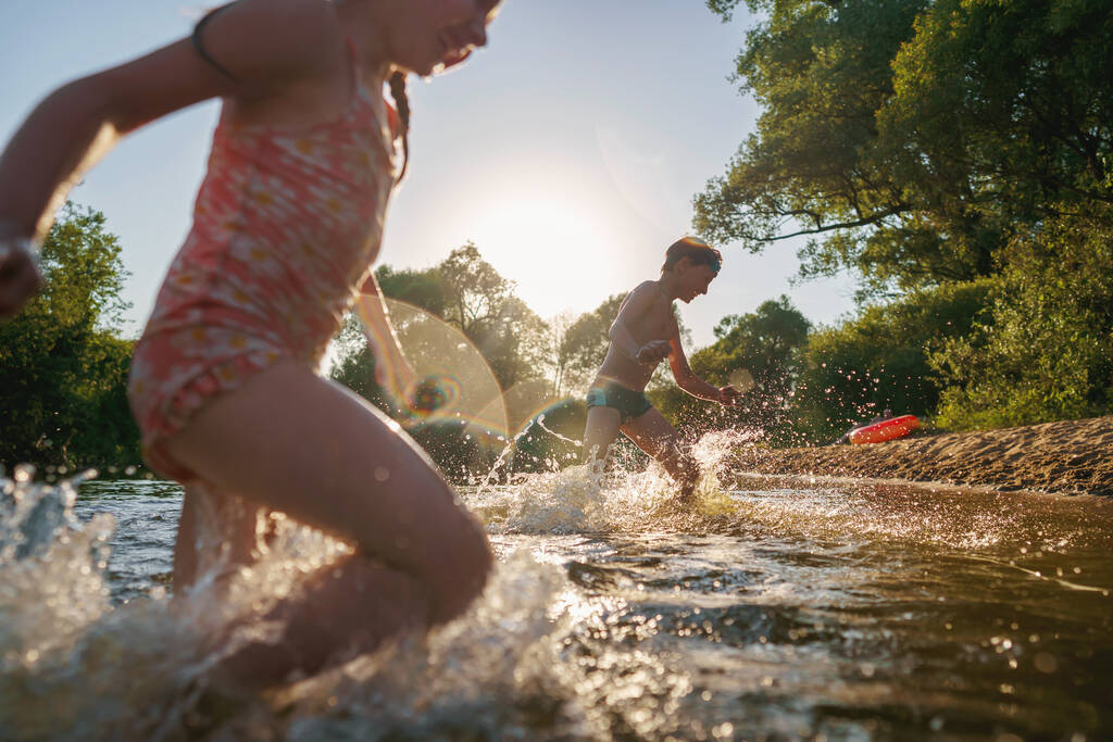 I bambini felici corrono dalla riva in acqua. Vacanze estive per bambini sulla riva di un lago o fiume. ragazzo e ragazza saltano in acqua, nuotano e schizzano in giro al tramonto. Vacanze attive. Immagine dinamica - Foto, immagini