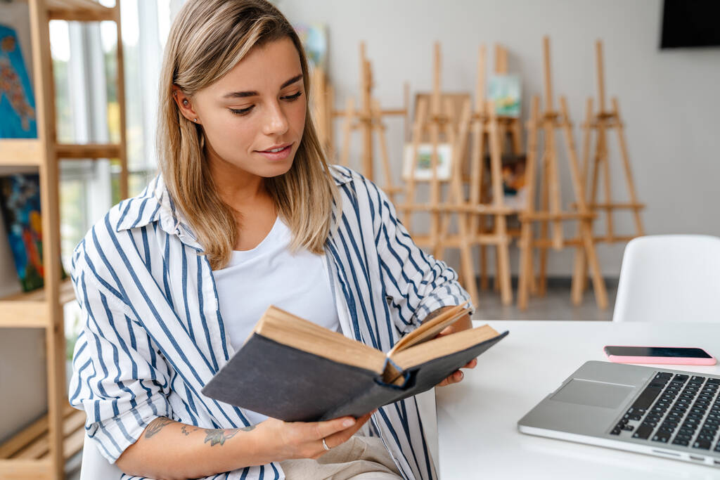 Usmívající se atraktivní mladá žena návrhářka čtení knihy, zatímco sedí v dílně - Fotografie, Obrázek