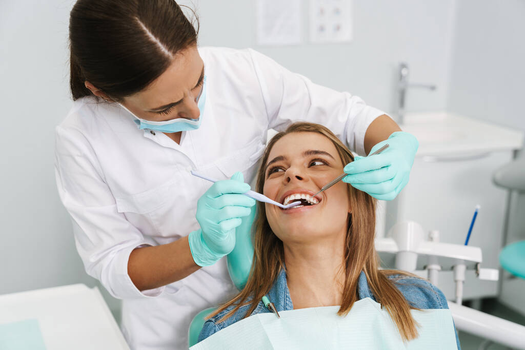 Mujer joven europea sentada en silla médica mientras la dentista fija sus dientes en la clínica dental - Foto, Imagen