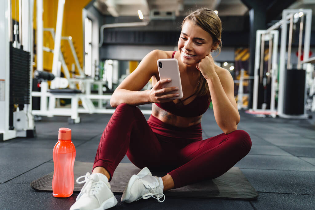 Happy athletic sportswoman using mobile phone and smiling while sitting on mat in gym - Photo, Image