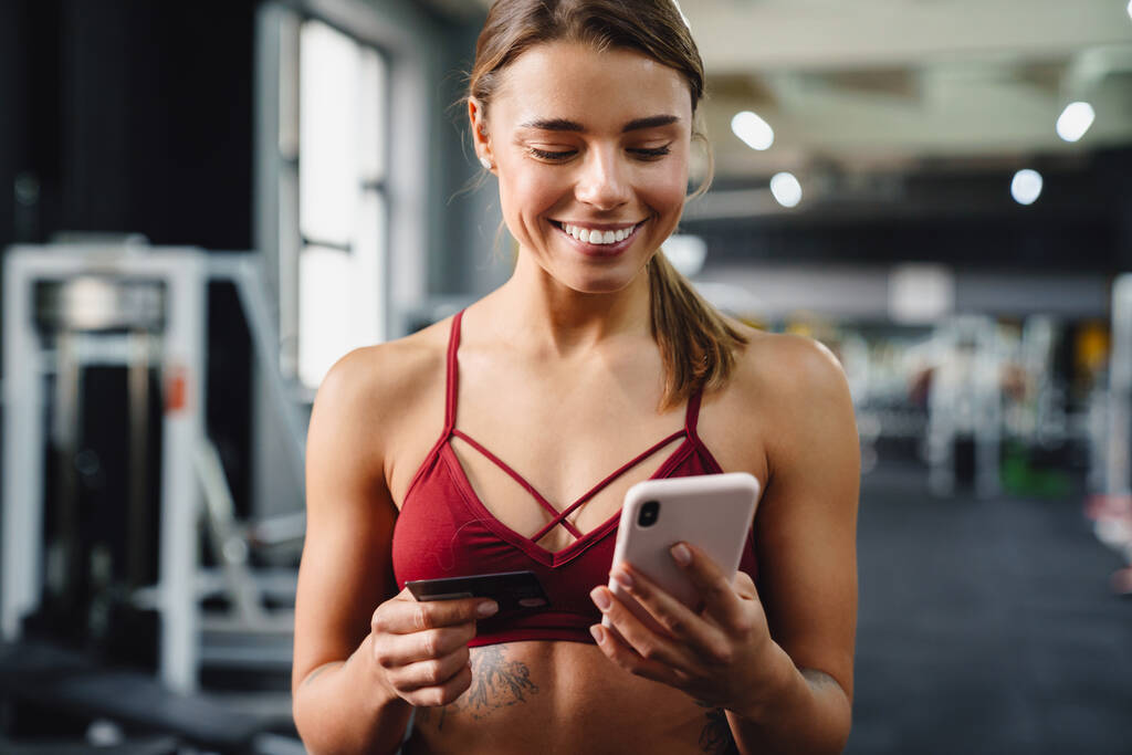 Zdjęcie uśmiechniętej młodej trenerki fitness korzystającej z telefonu komórkowego w siłowni trzymającej kartę kredytową - Zdjęcie, obraz