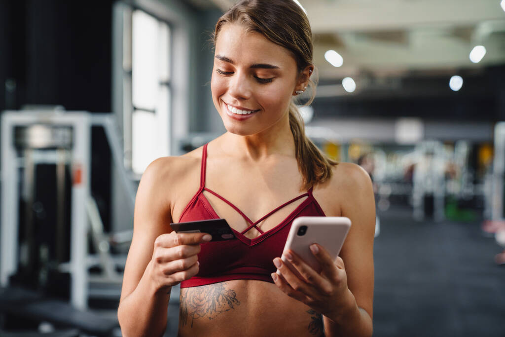 Photo of smiling young woman fitness coach using mobile phone in gym while holding credit card - Photo, Image