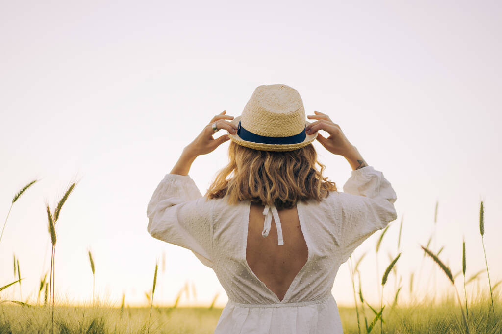 Sarı uzun saçlı, beyaz elbiseli, buğday tarlasında hasır şapkalı güzel bir kadın. Güneşte uçan saçlar, yaz. Hayalperestlerin zamanı, altın gün batımı. - Fotoğraf, Görsel