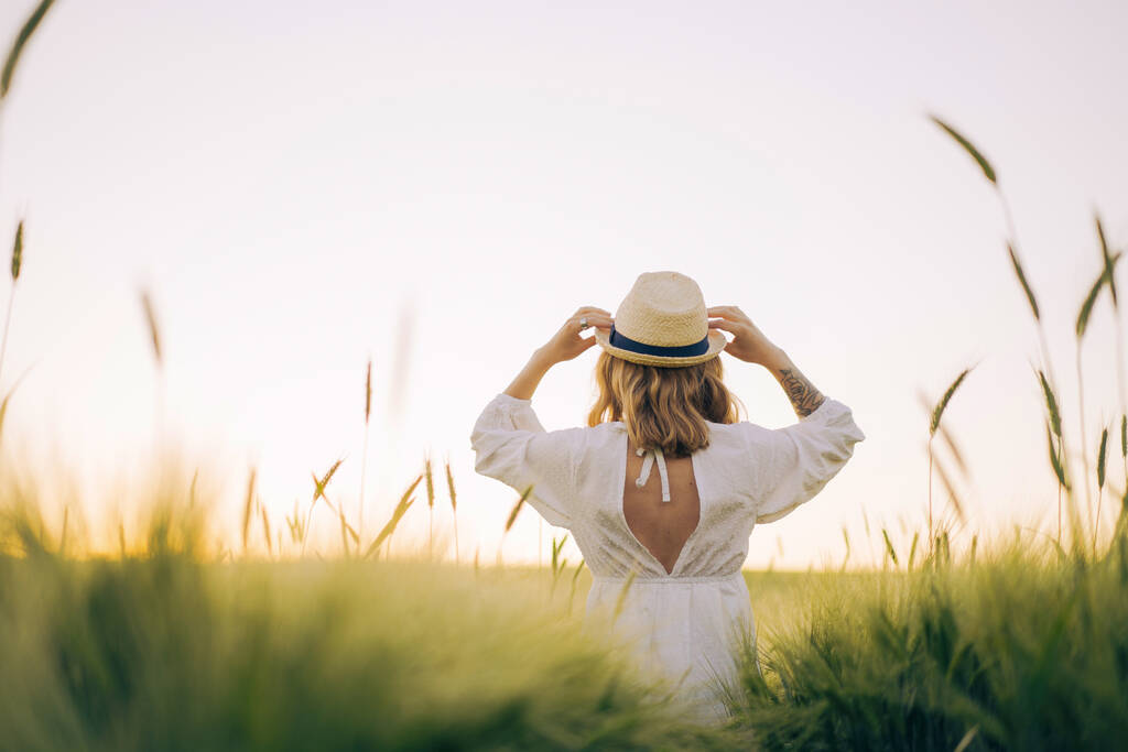 jonge mooie vrouw met blond lang haar in een witte jurk in een strohoed op een tarweveld. Vliegend haar in de zon, zomer. Tijd voor dromers, gouden zonsondergang. - Foto, afbeelding