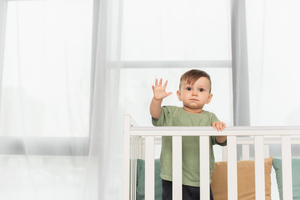 Kisgyermek fiú áll a babaágyban, és integet kezét otthon  - Fotó, kép