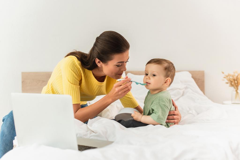 Mãe alimentando filho perto de laptop borrado na cama  - Foto, Imagem