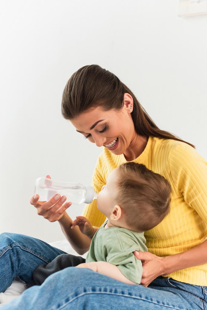 Mosolygó anya kezében cumisüveg, míg a fiú iszik a hálószobában  - Fotó, kép