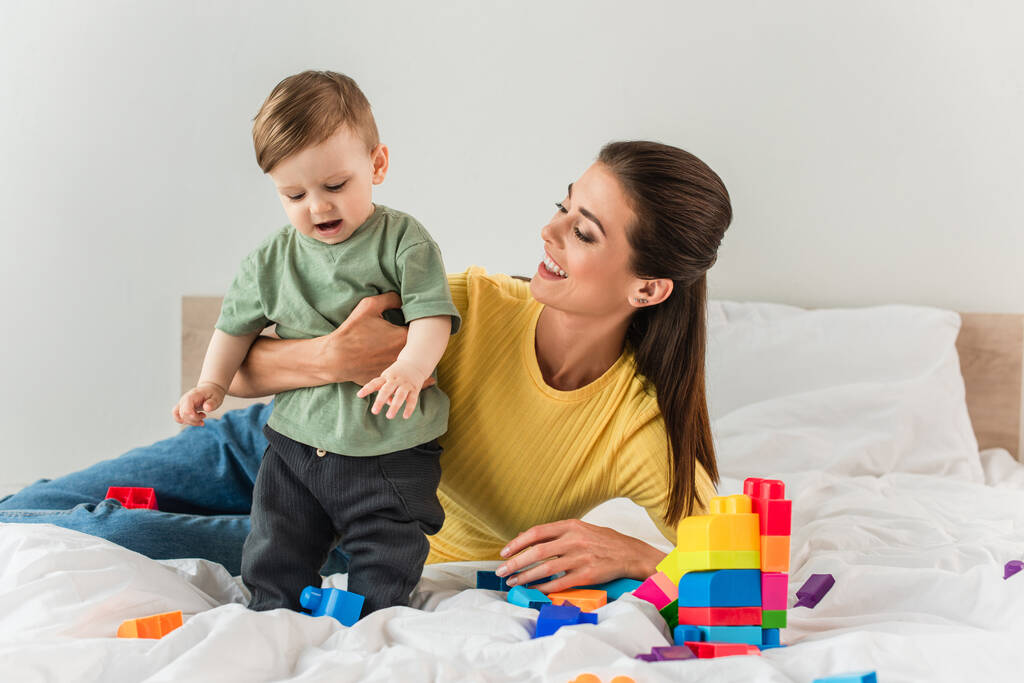 Happy mother embracing toddler son near building blocks on bed  - Photo, Image