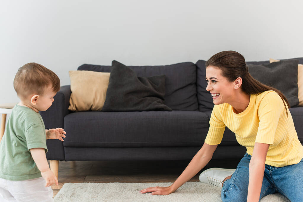 Vista lateral de la madre feliz mirando al niño en la sala de estar  - Foto, Imagen