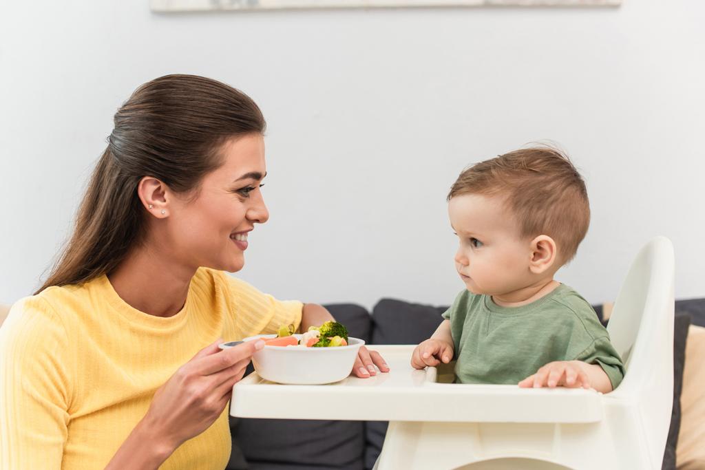 Side view of smiling mother holding bowl of vegetables near toddler son on high chair  - Photo, Image