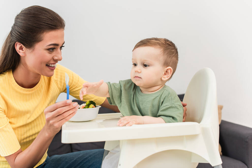 Улыбающаяся мать держит ложку рядом с ребенком малыша и овощи в миске дома  - Фото, изображение