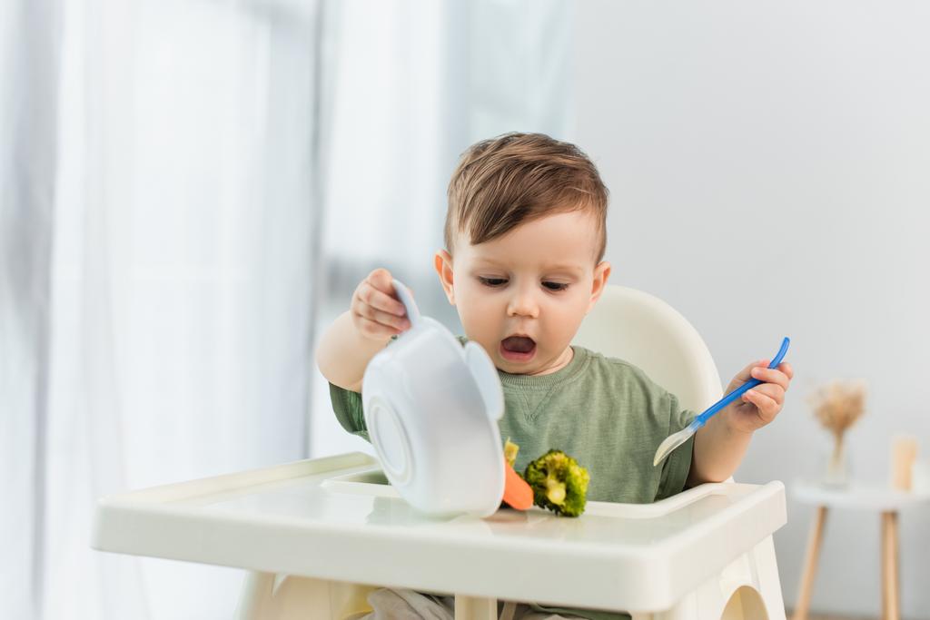 Дитина тримає ложку і поливає овочі на високому стільці
  - Фото, зображення