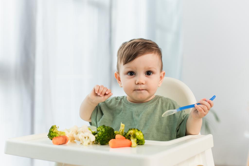 Kleinkind hält Löffel in der Hand und blickt in die Kamera neben Gemüse auf dem Hochstuhl  - Foto, Bild