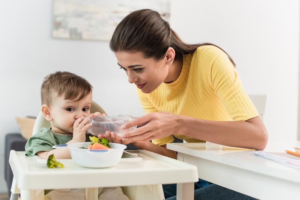 Kleinkind trinkt Wasser in der Nähe der Mutter und Nahrung auf Hochstuhl  - Foto, Bild