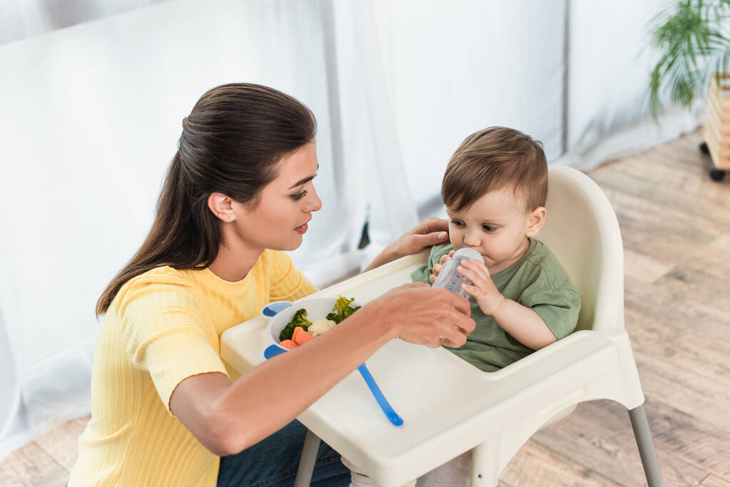 Madre sosteniendo biberón con chupete cerca del niño y tazón de verduras en la silla alta  - Foto, Imagen