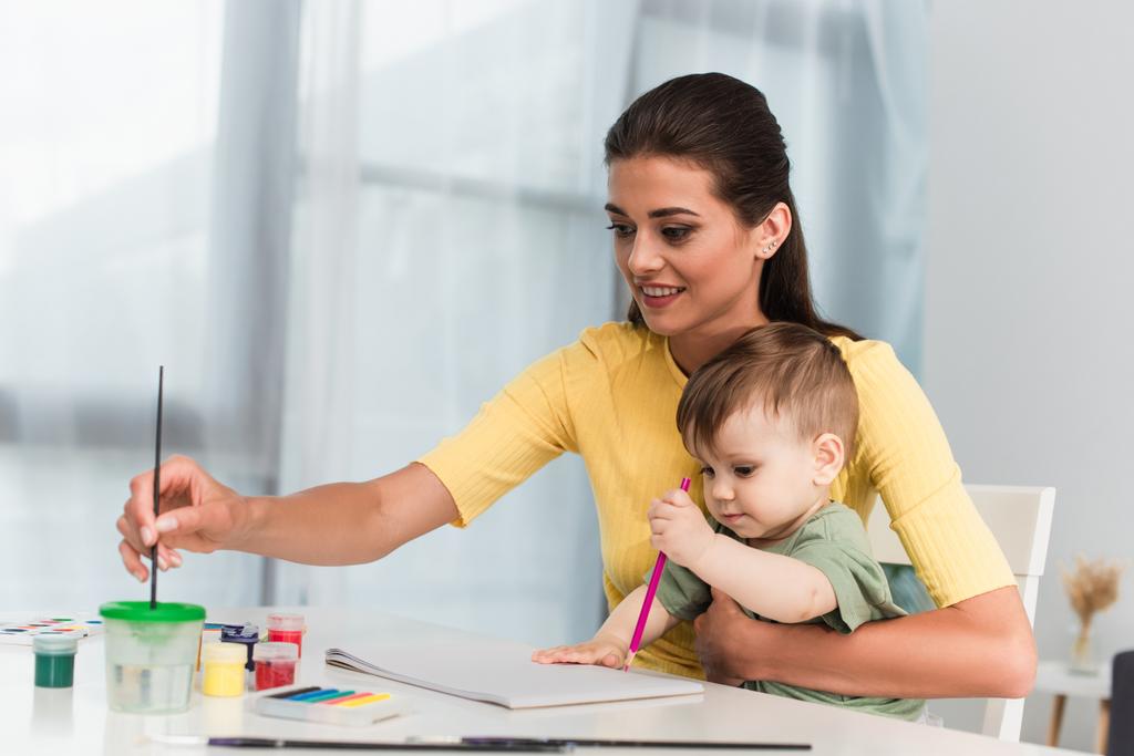 Mujer sonriente sosteniendo pincel de pintura cerca de niño con lápiz  - Foto, imagen
