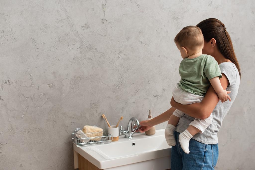 Robinet d'ouverture mère du robinet tout en tenant enfant en bas âge dans la salle de bain  - Photo, image