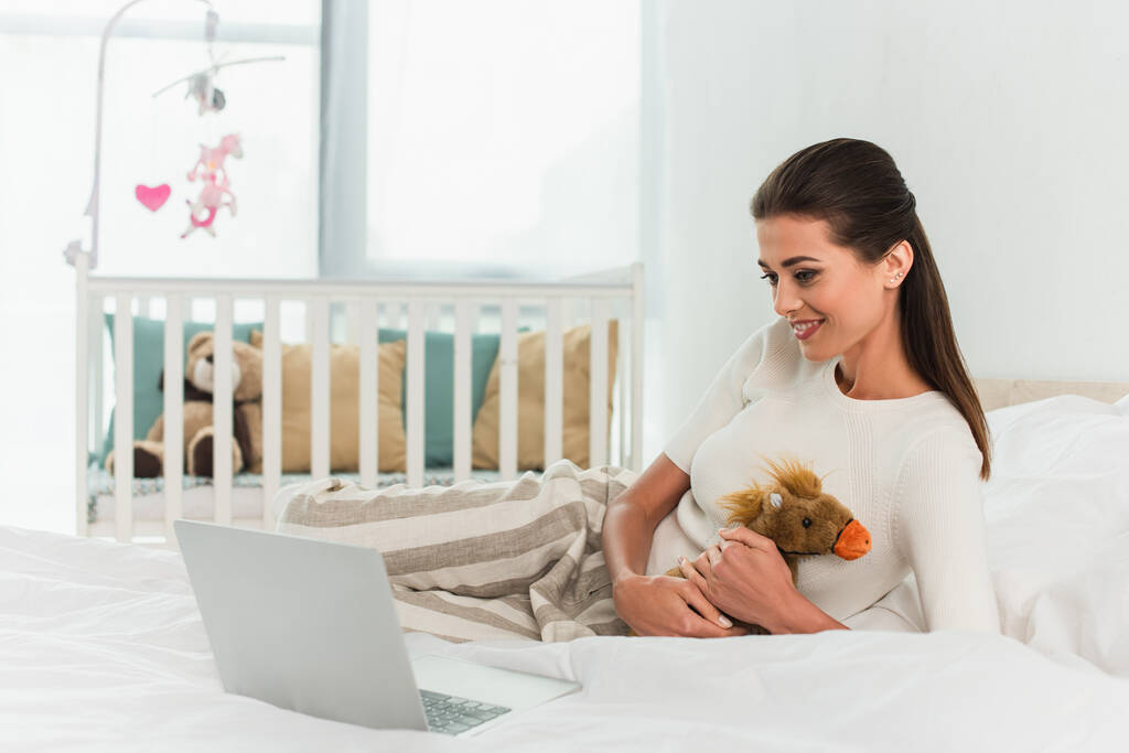 Lächelnde Frau hält Spielzeug neben Laptop auf Bett  - Foto, Bild