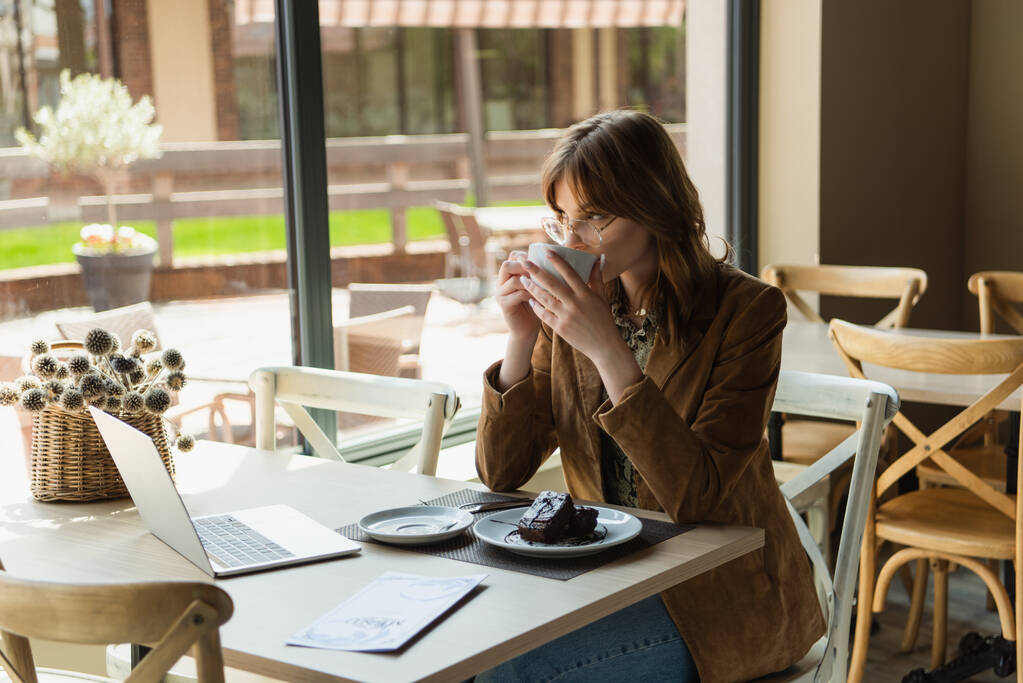 ノートパソコンの近くでコーヒーを飲んでいるスタイリッシュな女性、カフェでのデザートとメニュー  - 写真・画像