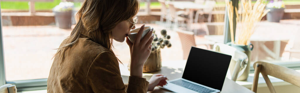 Πλευρική άποψη της νεαρής γυναίκας dinking καφέ κοντά θολή φορητό υπολογιστή στο καφέ, πανό  - Φωτογραφία, εικόνα