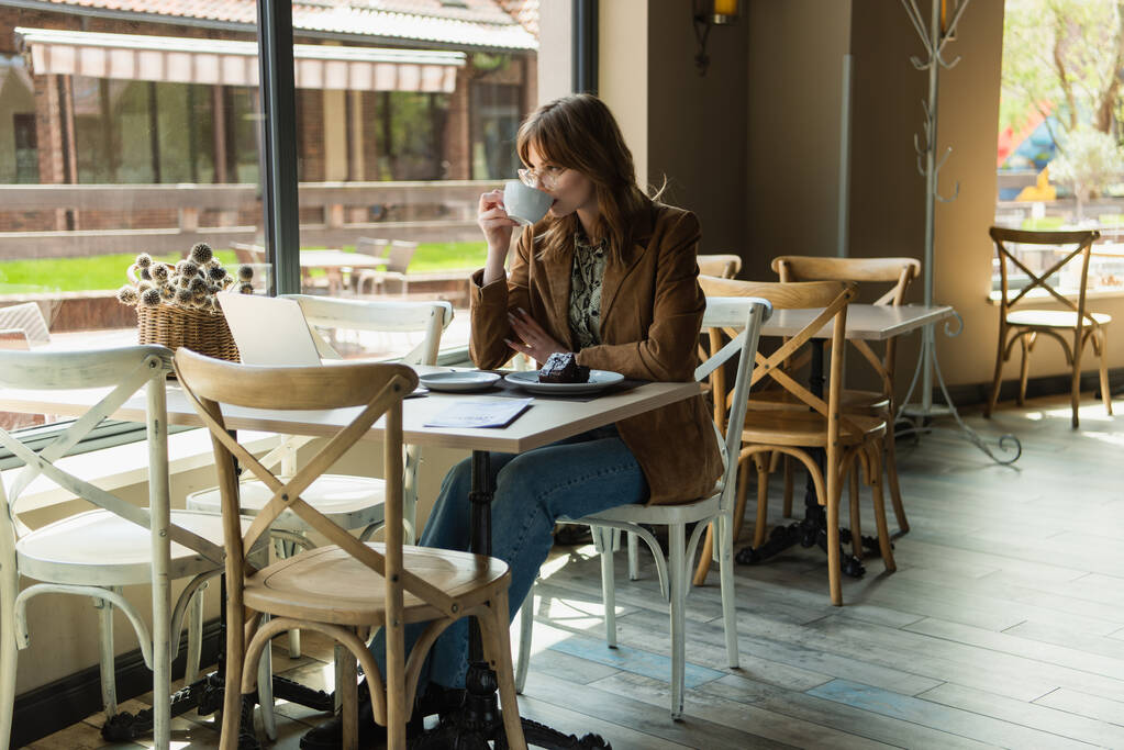 Stilvolle Frau trinkt Kaffee in der Nähe von Laptop und Dessert auf Tisch im Café  - Foto, Bild