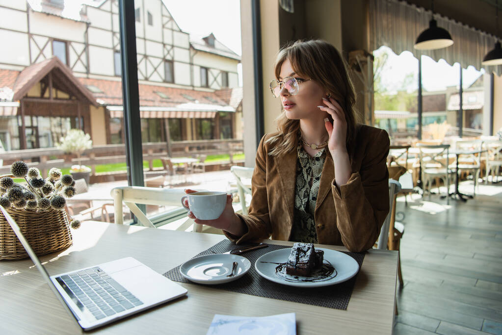 Módní žena v brýlích držící šálek v blízkosti chutného dezertu a notebooku v kavárně  - Fotografie, Obrázek