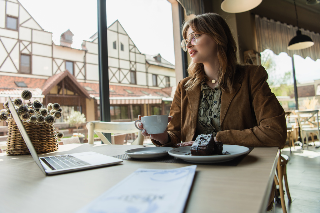 Mulher na moda segurando copo perto de sobremesa, laptop e menu borrado no café  - Foto, Imagem