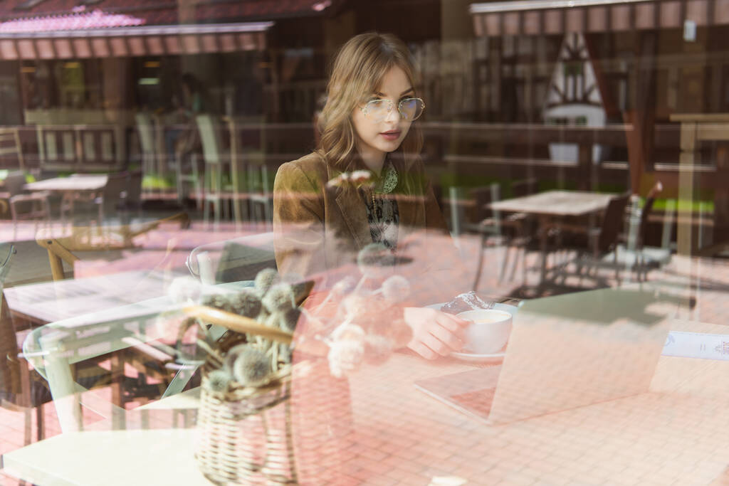 Ελεύθερος επαγγελματίας με καφέ και φορητό υπολογιστή κάθεται κοντά στο παράθυρο στο καφέ  - Φωτογραφία, εικόνα