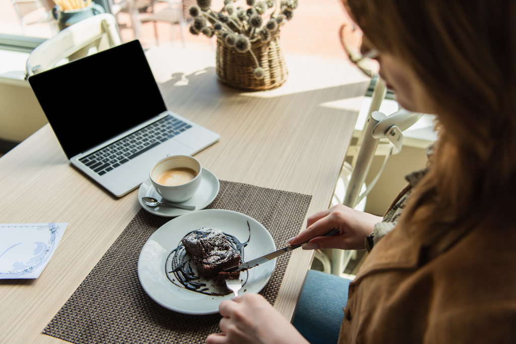 Размытая женщина режет десерт возле чашки кофе и ноутбука с пустым экраном в кафе  - Фото, изображение